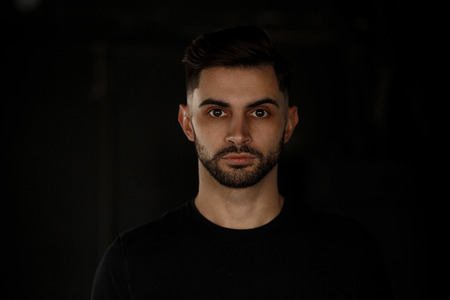 Close-up portrait of an attractive young handsome man with a stylish beard standing on a dark background, dressed in a black t-shirt.の写真素材