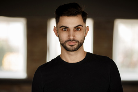 Portrait of a handsome young stylish man with a beard and hairstyle in a black t-shirt in the studio against dark backgroundの写真素材