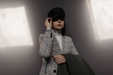 Beautiful stylish young model woman with hat and vintage fashion suite poses near a dark wall with light rays.の写真素材