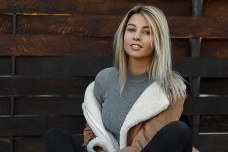 Pretty young American woman with a smile in a fashionable jacket with a gray knitted sweater near a wooden fence. Happy girl in the countrysideの写真素材
