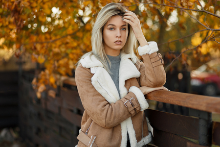 Autumn portrait of a pretty mysterious blonde girl in a stylish oversized beige jacket, resting in a city park near a wooden wall on the background of yellow leaves. A good day. Fashionable woman.の写真素材