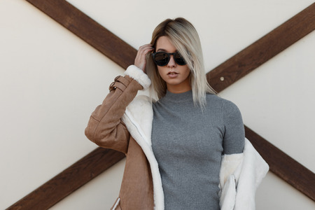 Young beautiful hipster girl with stylish sunglasses in fashionable gray vintage sweater and fashion jacket stands near a wall with wooden timber . Fashionable blond woman.の写真素材