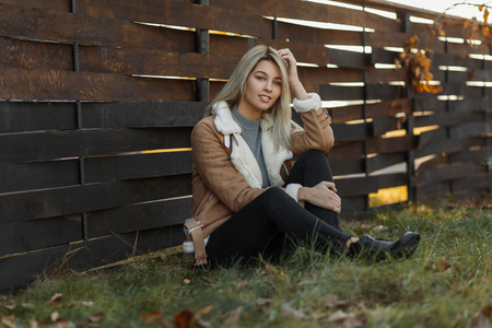 Happy cheerful woman in stylish beige jacket with fur with fashionable black jeans sits on green grass on the nature against the background of a wooden fence at sunset. Positive happy girl emotionsの写真素材