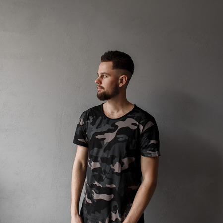 Attractive young man with a stylish hairstyle and brutal beard in a modern military green camouflage T-shirt posing  on a gray background. Fashionable modern guyの写真素材
