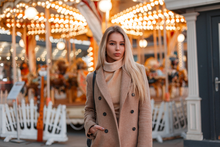 Young woman in a stylish warm autumn coat and a vintage knitted sweater stands in an amusement park in the city. Elegant fashion model girl.の写真素材