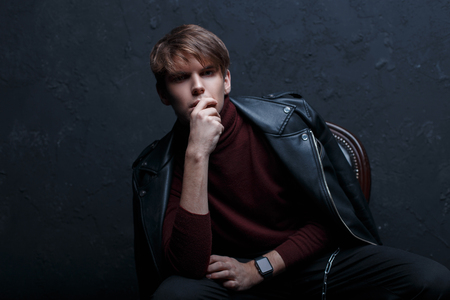 Young attractive man in a stylish leather black jacket in red golf in black jeans in trendy shoes with stylish watches posing in a dark studio near a black wall. Handsome stylish modern guyの写真素材
