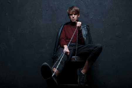 Young stylish man in a jacket in a vintage burgundy golf in trendy jeans and sneakers with a metal chain sits on a chair and looks into the camera in the studio against the black wall. Handsome guyの写真素材