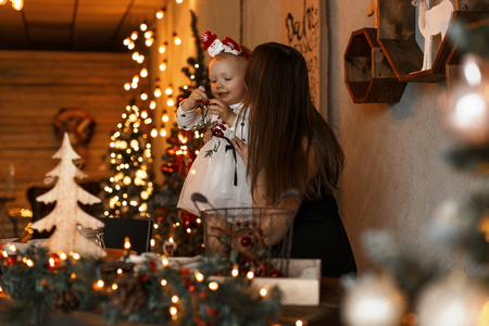 Pretty young mother in a black dress kissing a cute joyful little kid in a white dress in a decorated studio on the background of New Year's garlands. Christmas family holidayの写真素材