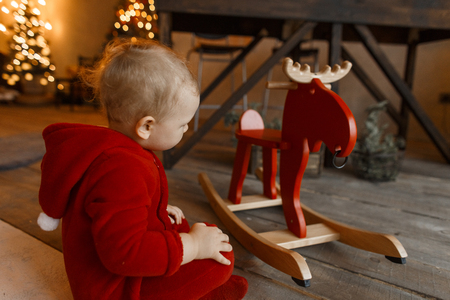 Little cute child in Christmas red plush fashionable clothes with a hood in a room decorated with festive garlands and toys sits on a wooden floor and looks at a toy deer chair. Happy holidayの写真素材
