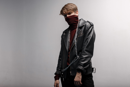 Cute young man wearing burgundy trendy golf in a black leather vintage jacket in black stylish jeans and metal chain, standing and posing indoors against a white wall. Stylish handsome guy.の写真素材