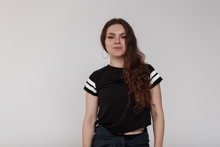 A pretty attractive young woman with brown curly hair in a fashionable black t-shirt with white stripes is standing in a bright room near a white background. Cute stylish girl smilingの写真素材