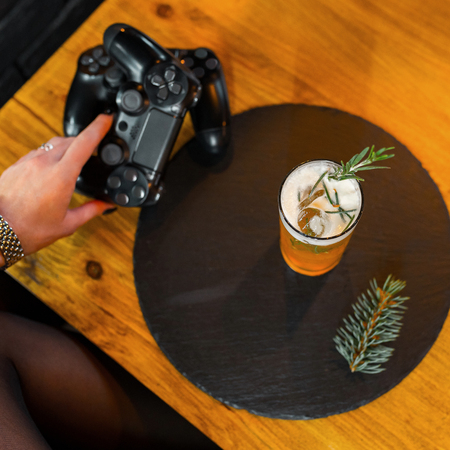 Young woman holds in her hand black modern gaming joysticks for playing video game against the background of a wooden table and an alcoholic cocktail. Modern video gamesの写真素材