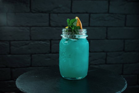 Tropical bright blue sweet cocktail the Blue Lagoon in a glass glass with pieces of ice decorated with mint leaves and orange slices stands on the table in the bar. Club nightlife. Good weekendの写真素材