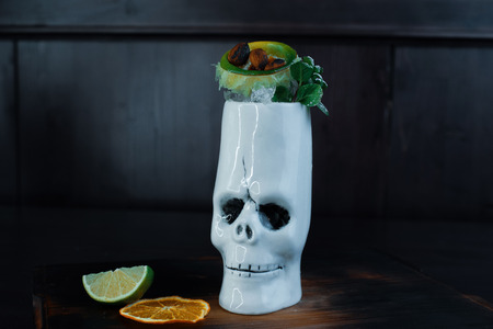 Tropical sweet cocktail with: tequila, white rum, fruit pieces and mint leaves on a wooden table in the bar. Alcoholic beautiful delicious cocktail in the original white skull glass.の写真素材