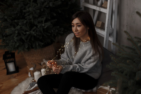 Pretty young woman in a gray sweater sits on a wooden floor in the room and holds a bright garland against the background of the New Year tree. Beautiful girl dreams of a Christmas miracle.の写真素材