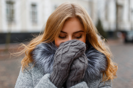 Charming funny young woman laughs and covers her face with her hands in knitted mittens on the background of vintage buildings. Cheerful nice girl.の写真素材