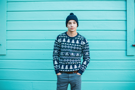 Young man with a beautiful smile in a knitted hat with blue eyes in a blue Christmas vintage sweater in jeans stands in the city near a bright blue wooden house. Charming guy.の写真素材