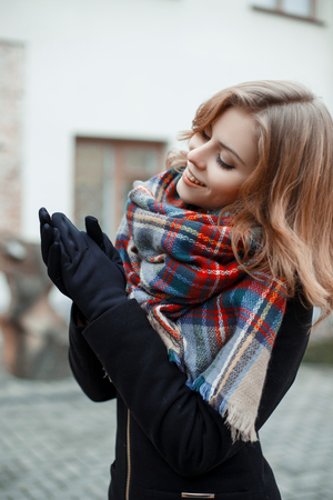 Wonderful attractive young woman in a stylish black coat with a woolen warm scarf in black gloves poses on the street autumn day on the background of the building. Beautiful girl smilingの写真素材