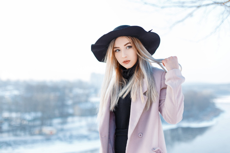 Sensual beautiful young woman with blond hair in a vintage hat in a winter pink coat in retro style is standing outside on the background of a river and snowy trees on a winter sunny day. Cute girl.の写真素材