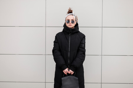Stylish hipster young woman in stylish sunglasses in a bandana in a fashionable long jacket with a leather stylish backpack near a white wall. American modern girl. Women's fashion.の販売画像