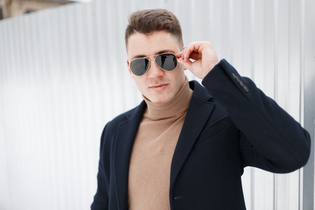 Porter of a stylish handsome young hipster male with a stylish hairstyle in a fashionable jacket in a knitted vintage sweater posing near a metal wall. American attractive guy on vacation.の写真素材