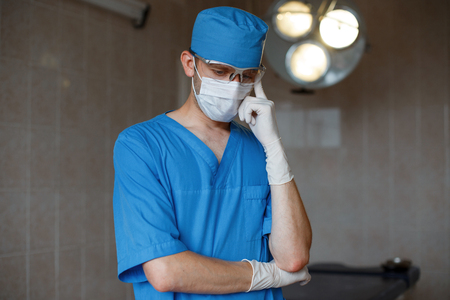 Young surgeon in a blue medical uniform in a protective white mask wearing rubber gloves is standing in the hospital and thinking. Neurosurgeon is preparing for surgery. Concept of health care.の写真素材