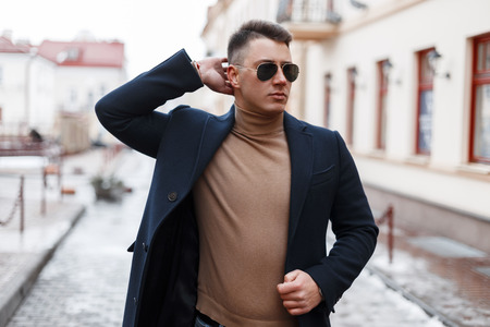 Handsome successful young hipster man in a vintage stylish coat in black sunglasses in a beige knitted sweater poses in the city in the warm day. Urban cute fashionable guy.の写真素材