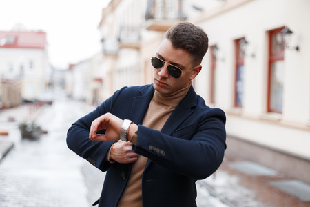 Stylish young businessman in an elegant coat in trendy sunglasses with a stylish hairstyle in a knitted sweater looks at a hand on the background of vintage buildings. Successful attractive man.の写真素材