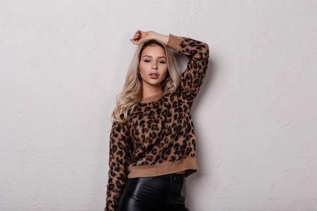 Young cute glamorous woman in a leopard vintage sweater in trendy black leather pants with curly hair poses indoors in a white room. Charming beautiful girl. Spring collection of women's clothing.の写真素材