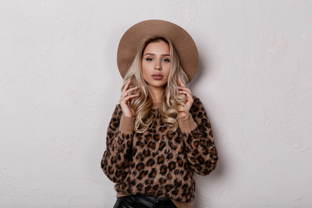 Beautiful charming young woman in a trendy stylish leopard sweater in trendy black leather pants and a beige hat posing indoors on a white background. Cute stylish blonde girl. Women's fashion.の写真素材