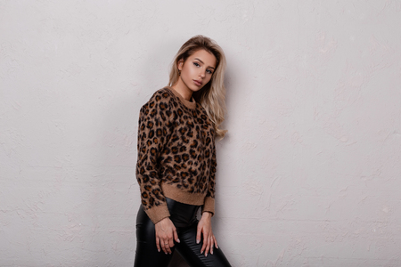 Charming pretty young woman in a stylish leopard jacket in black trendy leather pants is standing in a room near a white wall. Attractive blonde girl.の写真素材