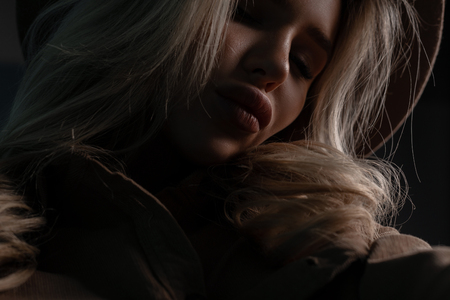 Close-up face of a young pretty woman with sexy lips with beautiful natural make-up with long disheveled blond hair in a dark background. Charming sweet girl with closed eyes relaxes.の写真素材