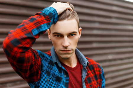 Young serious man with a stylish hairstyle in a trendy t-shirt in a fashionable plaid shirt poses near a vintage metal wall on a summer day. Stylish handsome guy. Portrait of a man's face.の写真素材