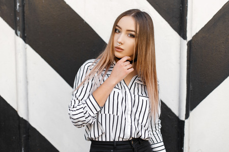 Beautiful young stylish woman in pierced nose in a stylish striped shirt in black jeans with a black choker around her neck poses near a black and white striped wall. Cute modern girl teenager.の写真素材