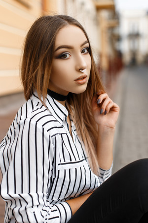 Beautiful attractive young woman in a velvet necklace with an earring in her nose in an elegant striped blouse in black jeans near a vintage building. Fashionable modern teenage girl outdoors.の写真素材