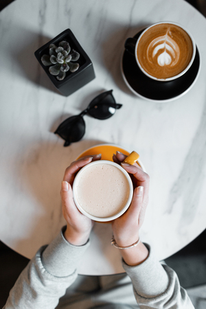Female hands holding a cup with hot coffee in the cafe. Young woman is sitting in the morning indoor. Top view of the table with coffee and sunglasses.の写真素材