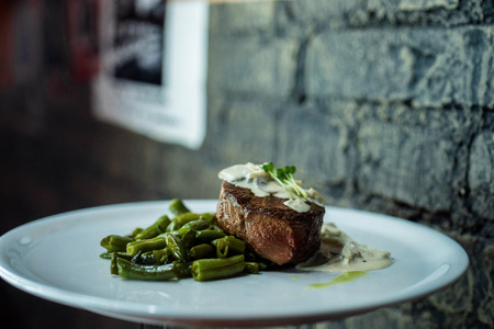 Hot appetizing tasty pork in mushroom sour cream sauce with baked green beans on a white plate in a bar on the background of a vintage wall. Lunch break.の写真素材