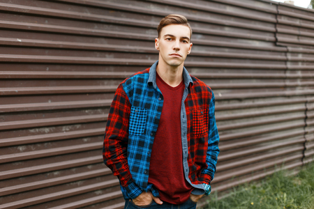 Stylish handsome young man in a fashionable red t-shirt in a trendy plaid multi-colored shirt in blue jeans outdoors near a vintage metal fence. Attractive American guy likes to go for a walk.の写真素材