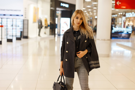 Pretty elegant young woman in a luxurious coat in a black shirt in vintage gray checkered pants with a leather handbag is standing in a modern shopping center.Cute fashion girl loves to make purchasesの写真素材