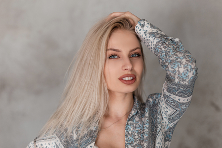Portrait of a pretty young blonde woman with blue eyes with plump sexy lips in a fashionable gray summer shirt with a pattern in a studio near the wall. Beautiful european girl. Female face portrait.の写真素材