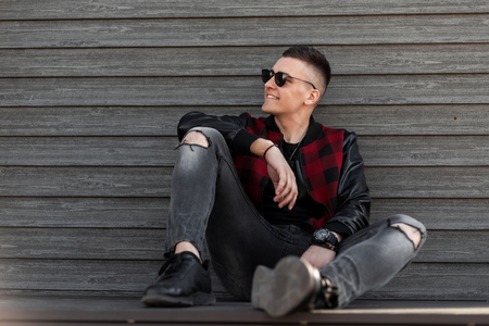 Happy american young hipster man in a fashionable checkered jacket with leather sleeves in ripped stylish jeans in sneakers sits and enjoys the weekend near a wooden wall. Handsome guy smiling.の写真素材