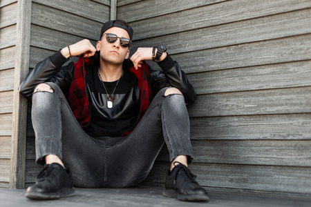American young hipster man in a black baseball cap in trendy sunglasses in a black t-shirt in a red checkered jacket resting sitting near a vintage wooden wall. Cool guy outdoors. Street style.の写真素材