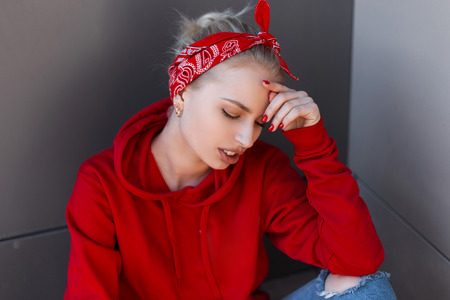 Beautiful european young blond woman in a red fashionable sweatshirt in a red stylish bandana with a hairstyle sitting outdoors on a sunny spring day. Pretty attractive girl enjoys the weekend.の写真素材