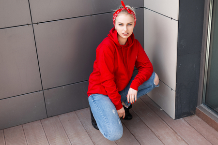 Young modern stylish woman in black boots in a red sweatshirt in ripped jeans with a fashionable bandana sitting outdoors near a gray wall. Beautiful girl enjoys the rest. Youth American style.の写真素材