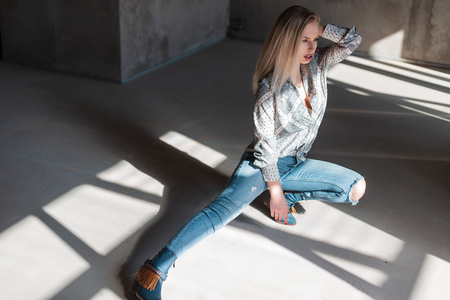 American young blonde woman in stylish shirt in vintage ripped jeans in trendy cowboy boots posing sitting indoors with sunlight. Sexy beautiful fashion model girl relaxes in a sunny room.の写真素材