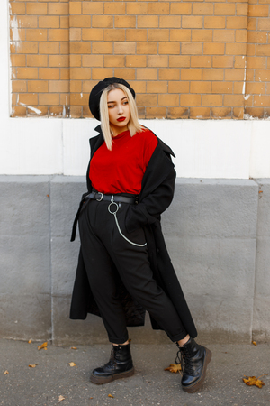 Pretty elegant young blond woman in a fashionable coat in trendy pants in a red shirt in a beret in leather boots is resting standing near a vintage brick wall on an autumn day. Cool girl outdoors.の写真素材