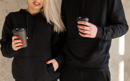 Young happy women blonde with a young stylish man with a beard in fashionable black clothes are standing near the wall indoors and holding coffee in hands. Coffee break. Close-up.の写真素材