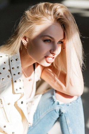 Portrait of a happy pretty young woman with beautiful blue eyes with blond hair with plump sexy lips in a leather fashionable spring jacket on a sunny day. Cute girl model enjoys bright sunlight.の写真素材