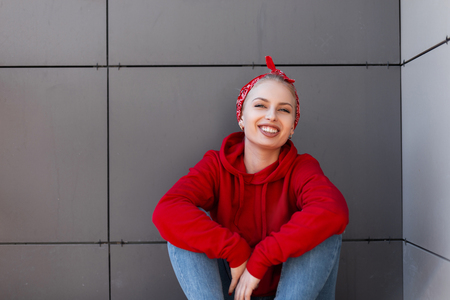 Cheerful young woman with a beautiful smile in a stylish red sweatshirt in a  trendy bandana in blue jeans is sitting on a sunny spring day near a gray building. Attractive girl model resting outdoorsの写真素材