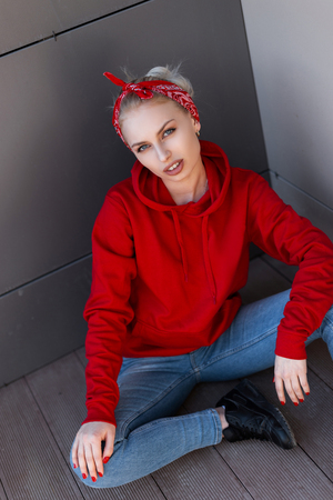 American modern young blond woman in a trendy red hooded sweatshirt in a red bandana in leather boots in blue jeans is sitting near a gray building on a summer day. Glamorous beautiful girl model.の写真素材